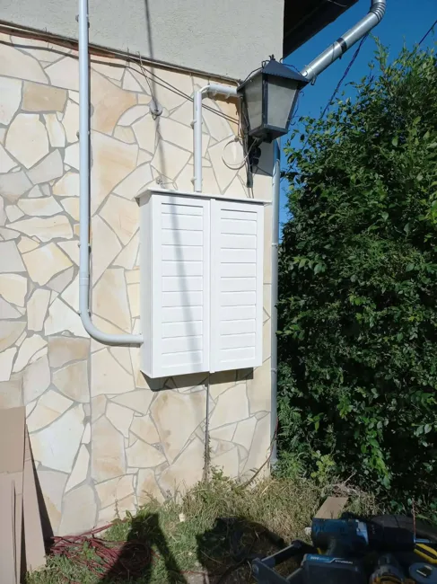 Fehér, kétajtós fa villanyóraszekrény-burkolat kőburkolatos külső falon.